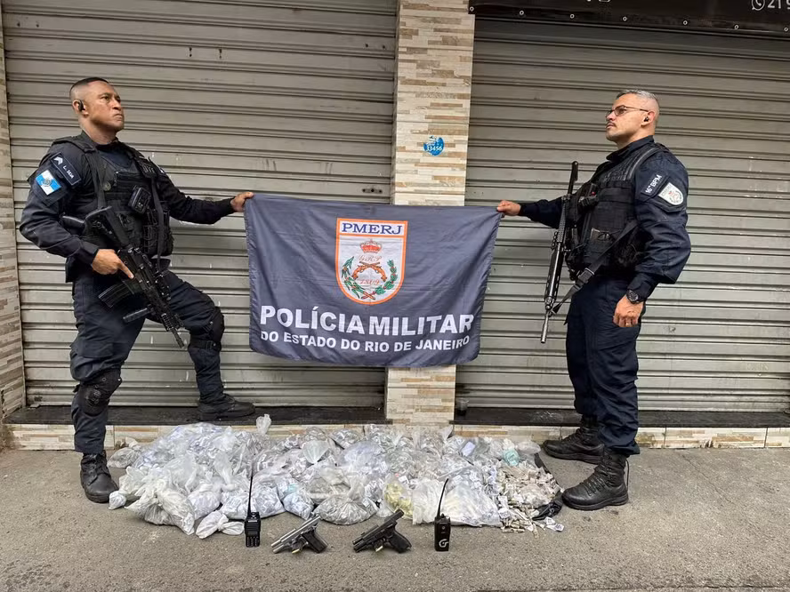 Policiais militarem ao lado de drogas, armas e rádios comunicadores apreendidos durante operação no Quitungo